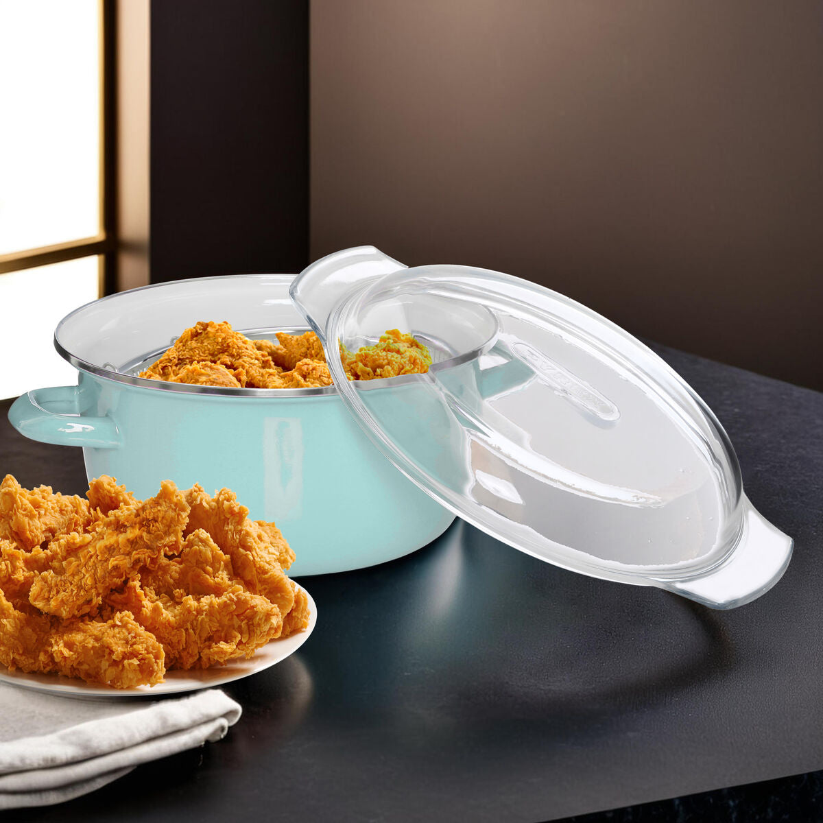Turquoise ceramic pot with a clear lid on a dark surface, accompanied by fried chicken on a plate.