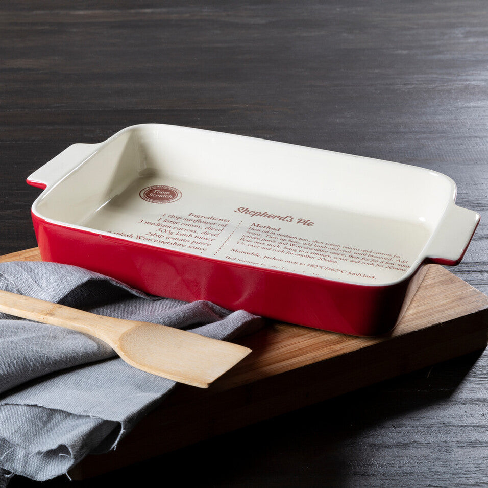 Red ceramic baking dish with white interior on a wooden board, accompanied by a wooden spatula and gray cloth.