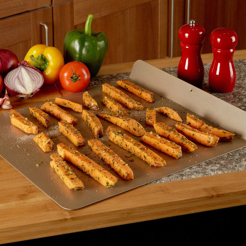 Baked sweet potato fries on a baking sheet with vegetables and spices on a wooden countertop.