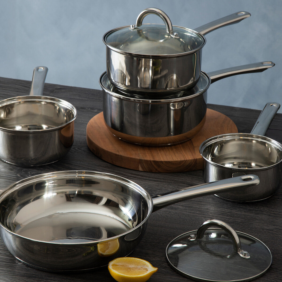 Set of stainless steel cookware including pots, pans, and lids on a wooden surface with a blue background.