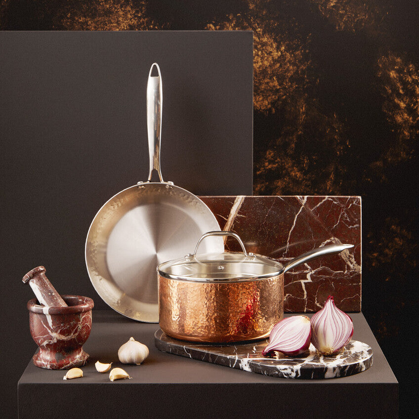 Copper cookware set on a dark surface with a marble cutting board and ingredients.