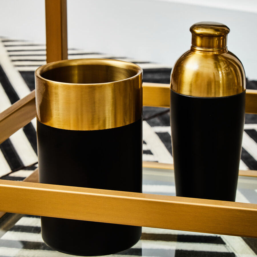 Two black and gold cylindrical containers on a glass table with a striped rug in the background.