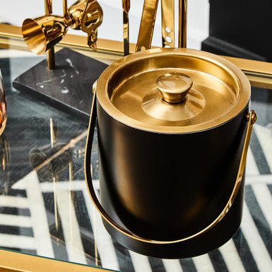Black and gold ice bucket on a reflective surface with decorative elements in the background