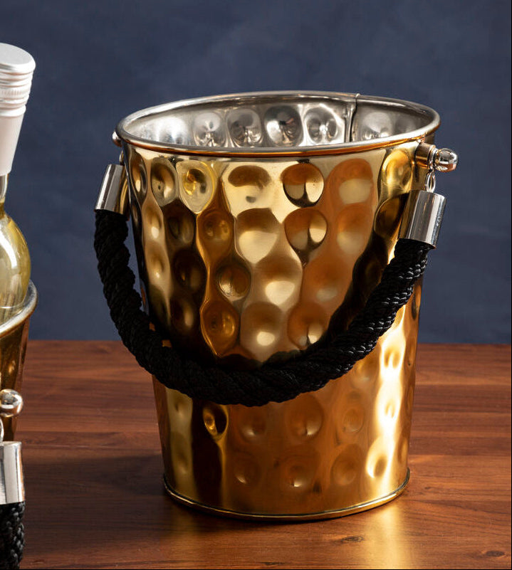 Gold ice bucket with black rope handle on a wooden surface next to a bottle of white wine.