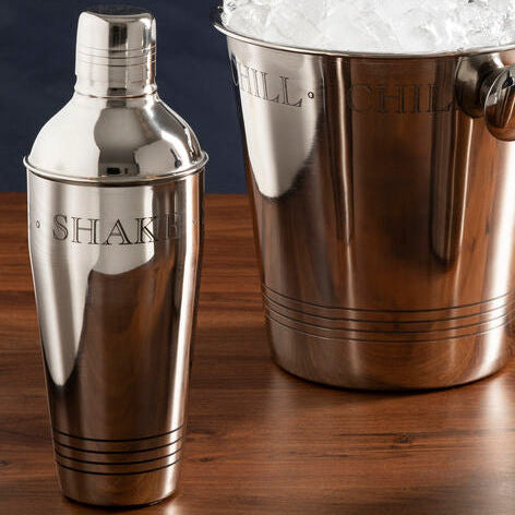 Metallic cocktail shaker, ice bucket with ice, and wine cooler on a wooden surface with a dark background.