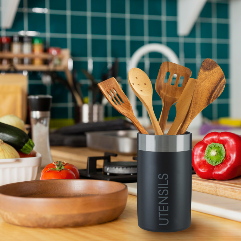 Black utensil holder with wooden spoons on a kitchen counter with vegetables and cookware.