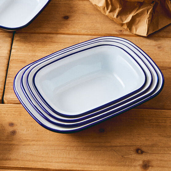 Set of white enameled cookware with blue rims on a wooden surface with eggs in a paper bag.