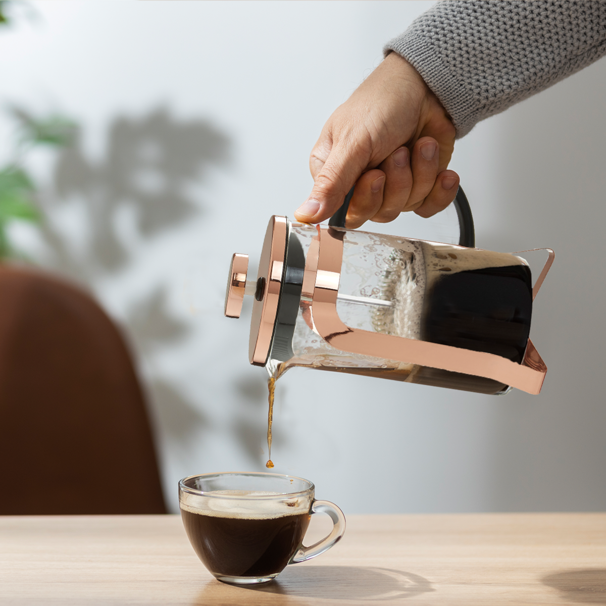 Person pouring coffee from a bronze French press into a clear glass mug on a wooden table.