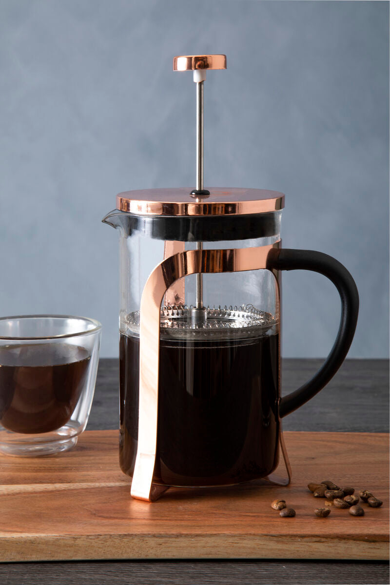 French press with copper accents on a wooden surface next to a glass of coffee and scattered coffee beans.