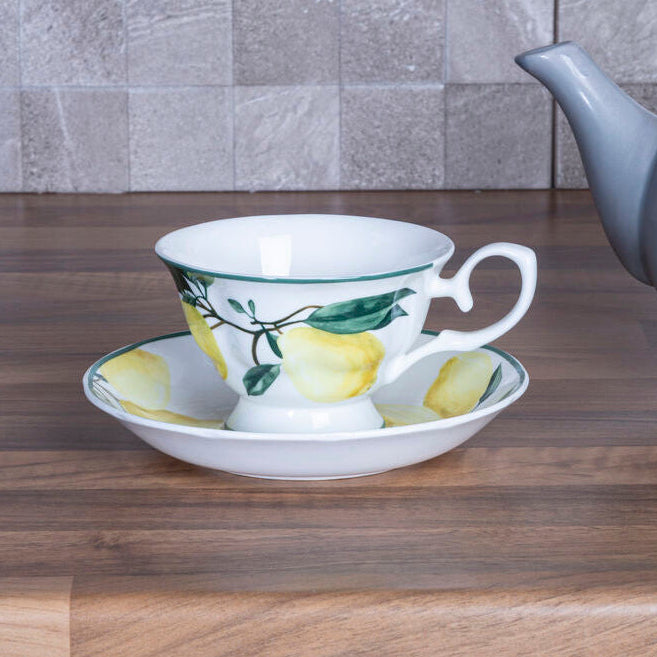 Teacup with lemon design on a saucer next to a gray teapot on a wooden surface with a tiled wall background.
