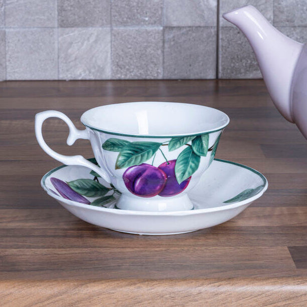 Teacup with floral design on a saucer next to a teapot on a wooden surface with a tiled wall background.