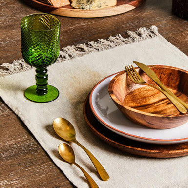 Table setting with wooden bowl, green glass, gold cutlery, and cheese platter.