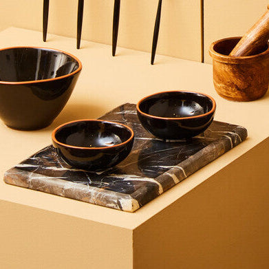 Set of black ceramic bowls, a mortar and pestle, and other kitchen items on beige shelves against a textured brown wall.