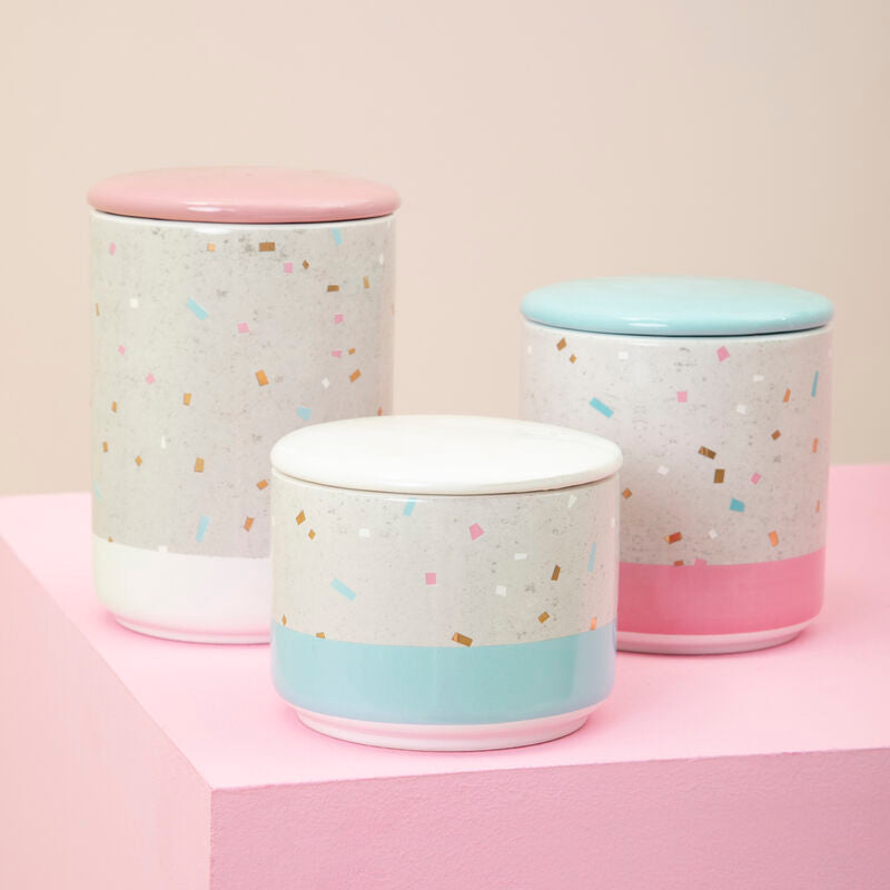 Three ceramic jars with colorful confetti pattern and pastel lids on a pink surface.