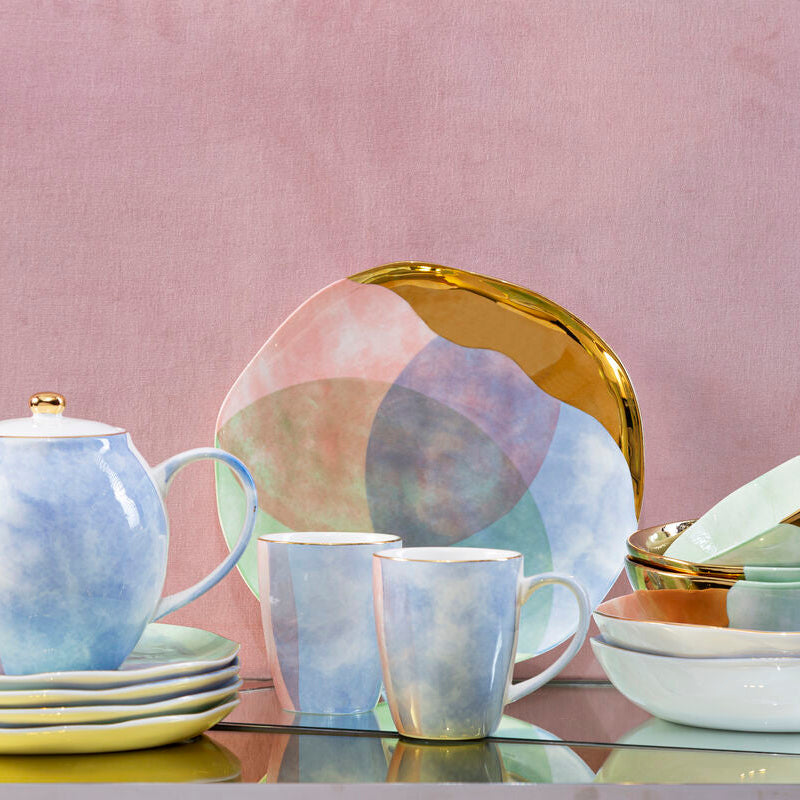 Ceramic tableware set with teapot, mugs, and plates on a reflective surface with a pink background