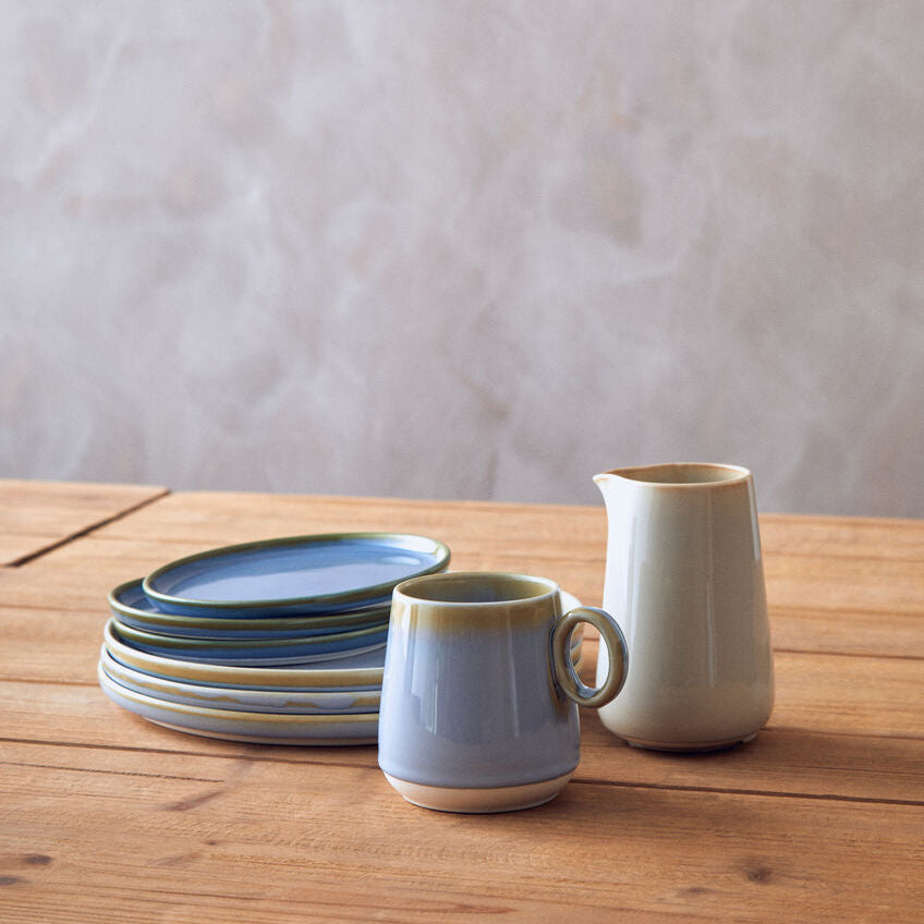 Ceramic tableware including a mug, plates, and a pitcher on a wooden surface with a neutral background.