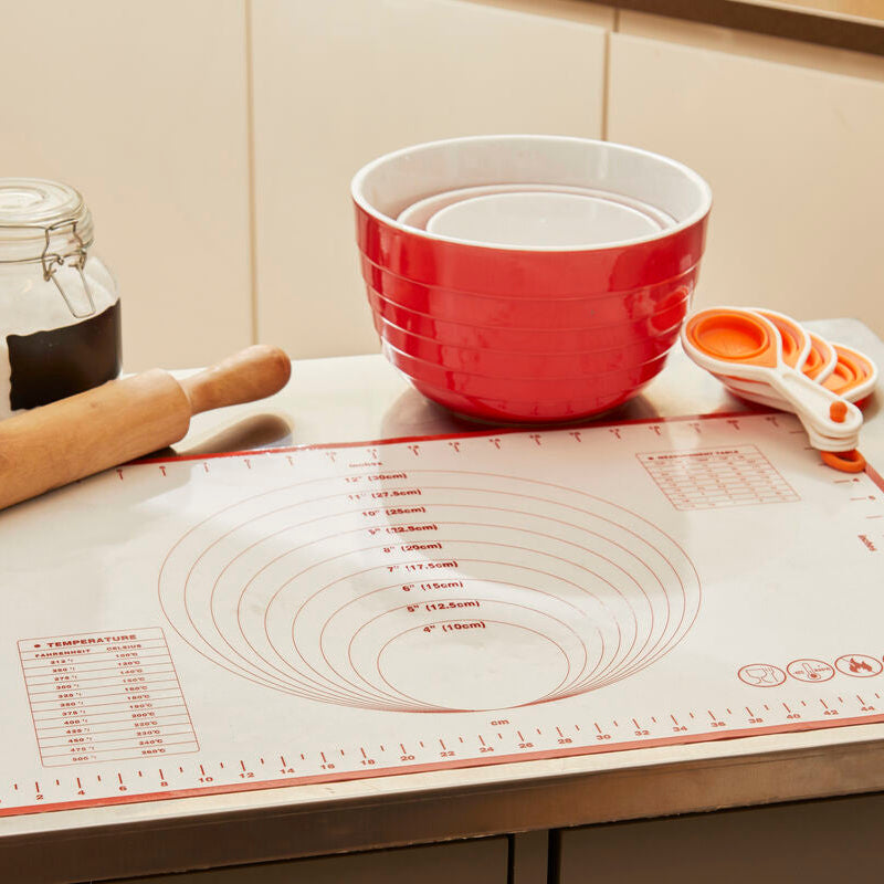 Baking tools including a red mixing bowl, rolling pin, and measuring cups on a kitchen counter with a silicone mat.