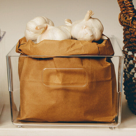 Clear container with brown fabric and garlic bulbs next to a wicker basket on a neutral background