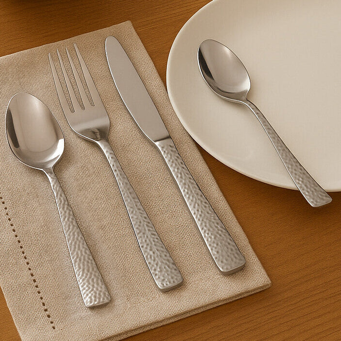 Silver cutlery set on a beige napkin with a white plate, bowl of salad, and glass of water on a wooden table.