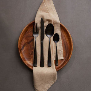 Set of black cutlery on a brown plate with a beige napkin against a gray background