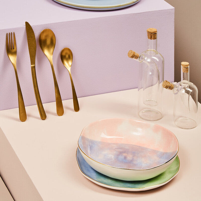 Dining table setting with ceramic bowls, gold cutlery, and glass bottles on a neutral background