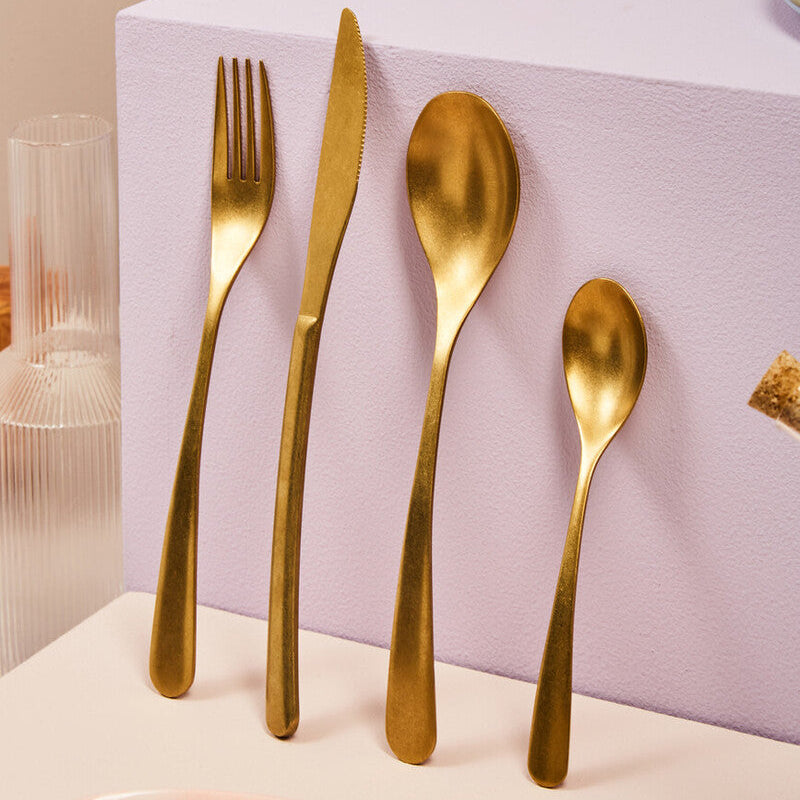 Set of gold cutlery including fork, knife, and two spoons on a pink surface with a blurred background.
