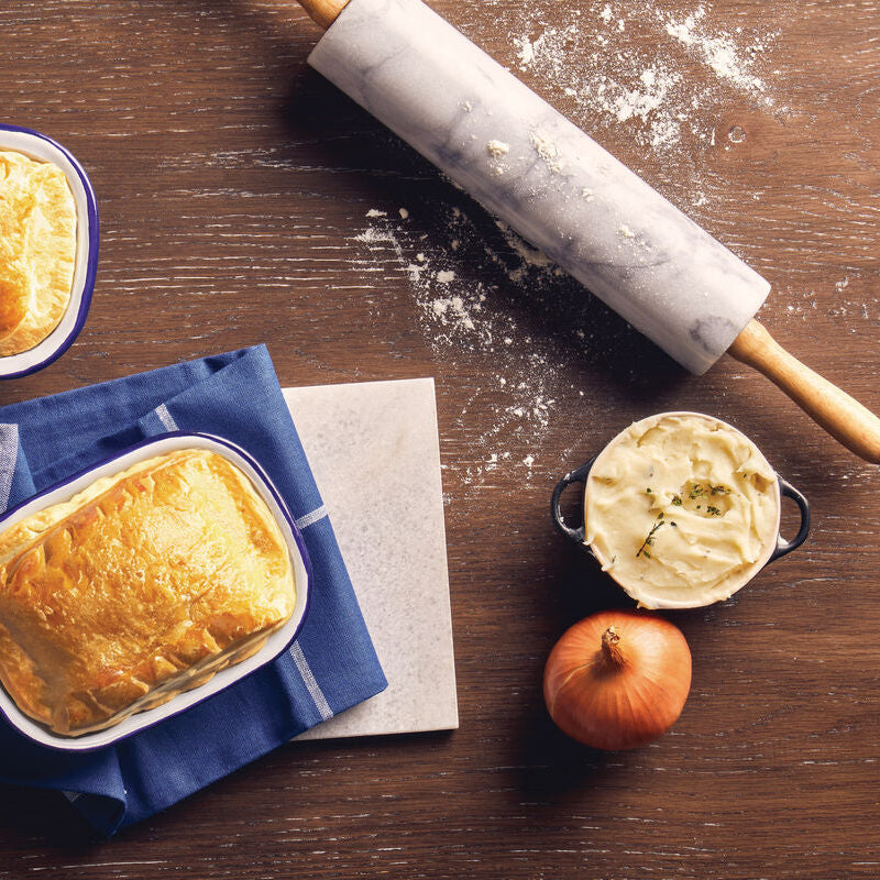 Two pies in baking dishes on a wooden table with a rolling pin and ingredients.