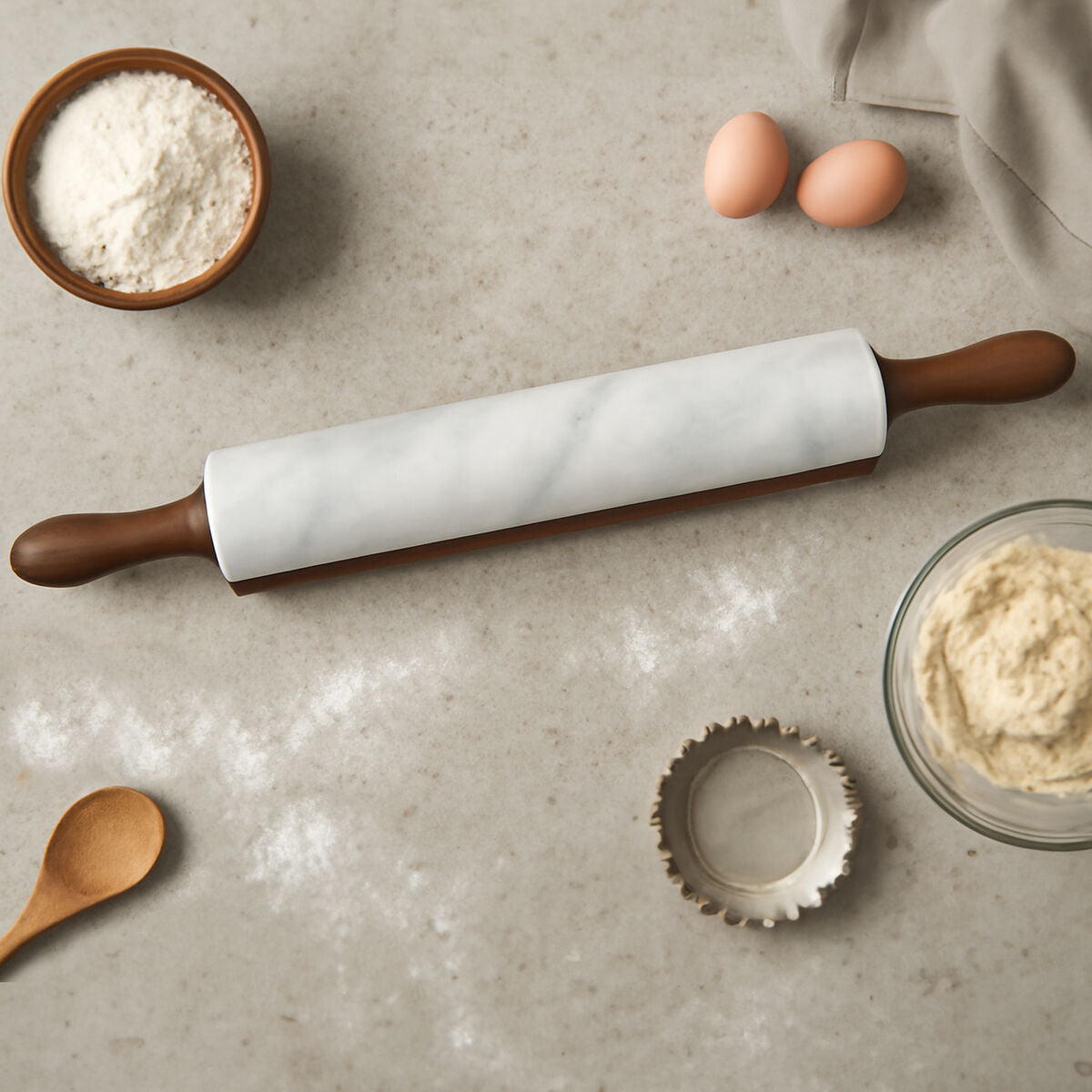 Rolling pin with white paper on a gray surface with flour, eggs, and dough.