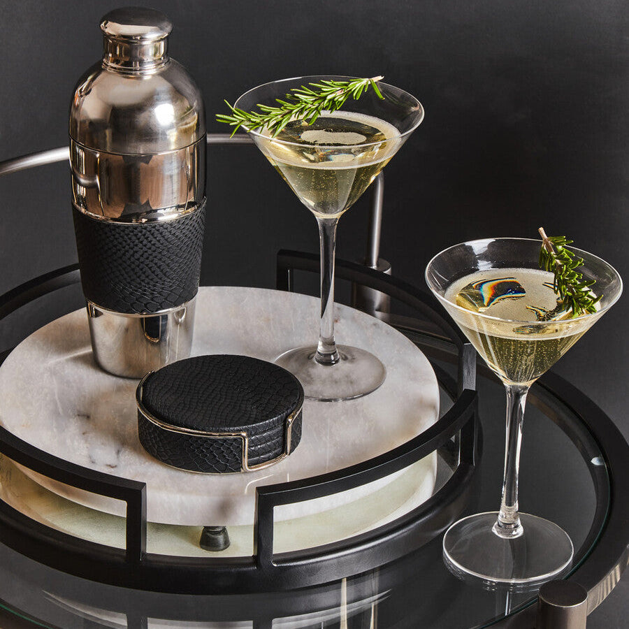Two cocktail glasses with a shaker on a round marble tray against a dark background