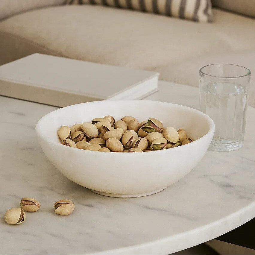 Bowl of pistachios on a marble table with a glass of water and a book in the background.