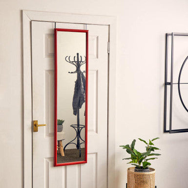Full-length mirror with red frame leaning against a white door in a room with a plant and rug.