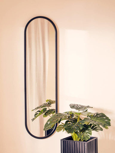 Oval mirror with black frame on a beige wall next to a potted plant.