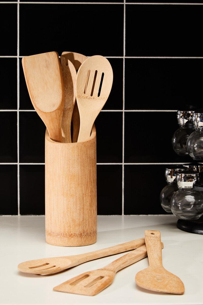 Set of wooden kitchen utensils in a cylindrical holder on a kitchen counter with black tiled wall.