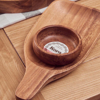 Wooden spoon-shaped dish with a small bowl on a wooden surface, featuring the Kora brand.