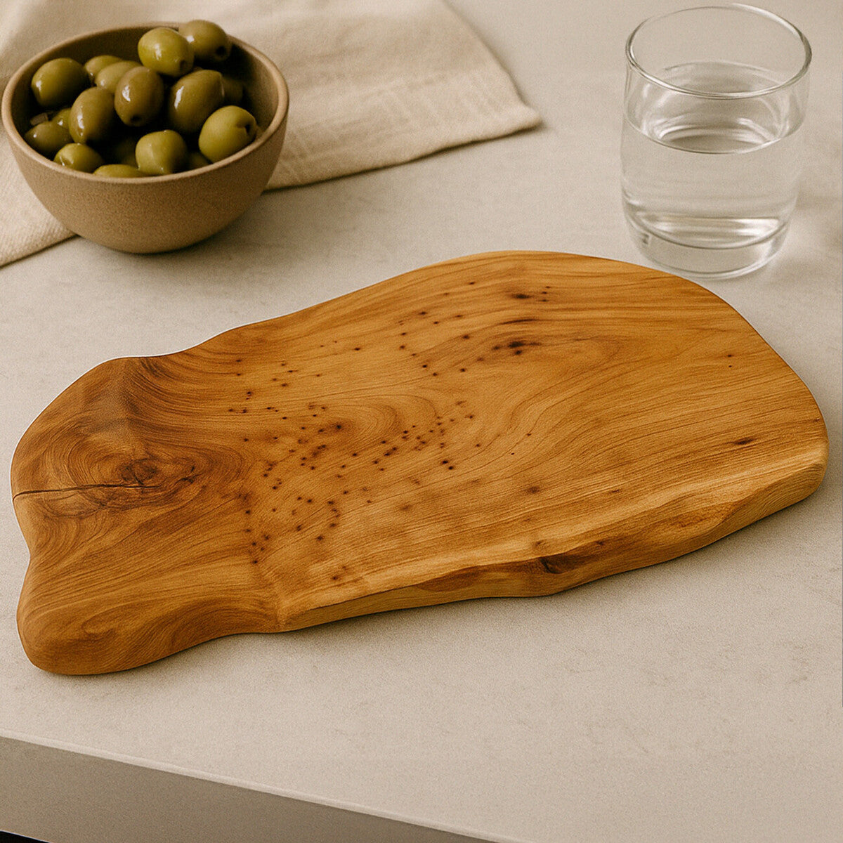 Wooden cutting board on a light surface with a bowl of olives and a glass of water.