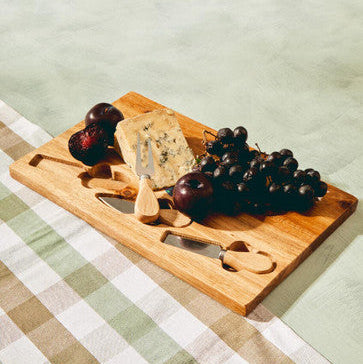 Wooden cutting board with cheese, grapes, and a knife on a checkered tablecloth.