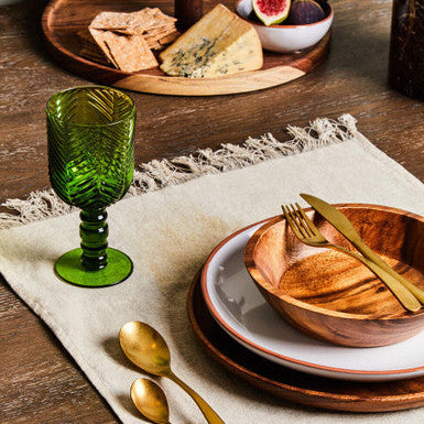 Table setting with wooden bowl, green glass, gold cutlery, and cheese platter.