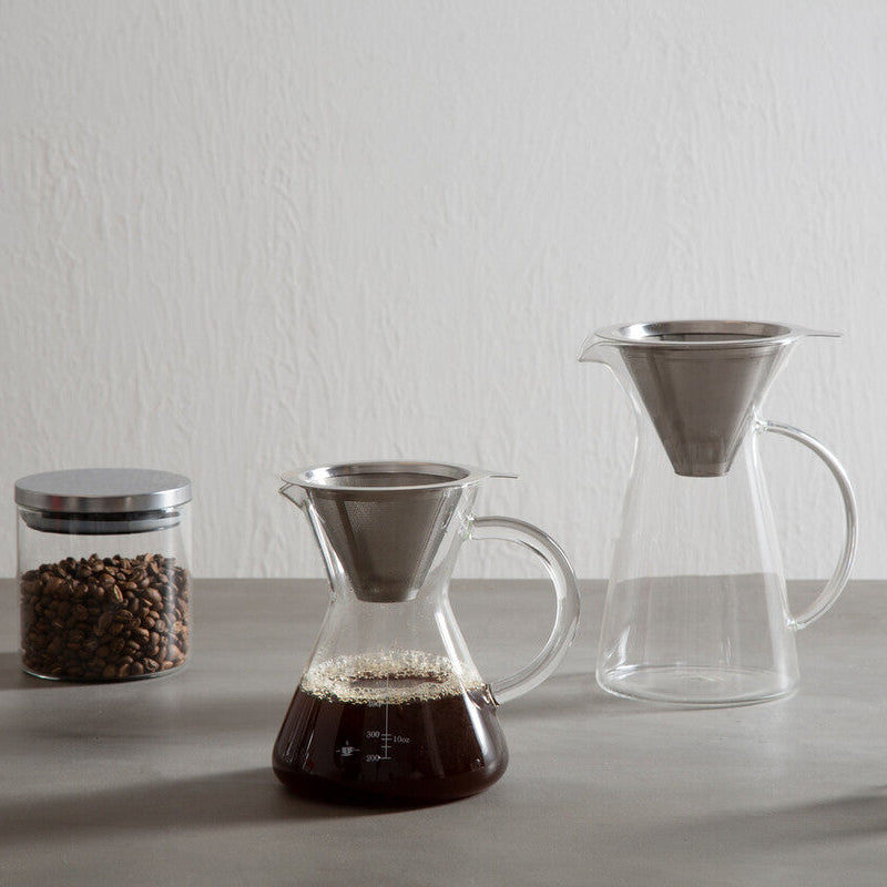 Coffee-making setup with coffee beans, coffee filter, glass carafe, and clear cup on a light gray surface.