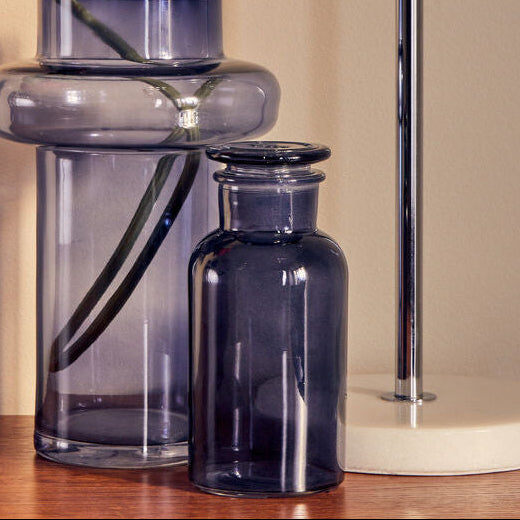 Glass vase with a pink flower on a wooden surface next to a lamp.