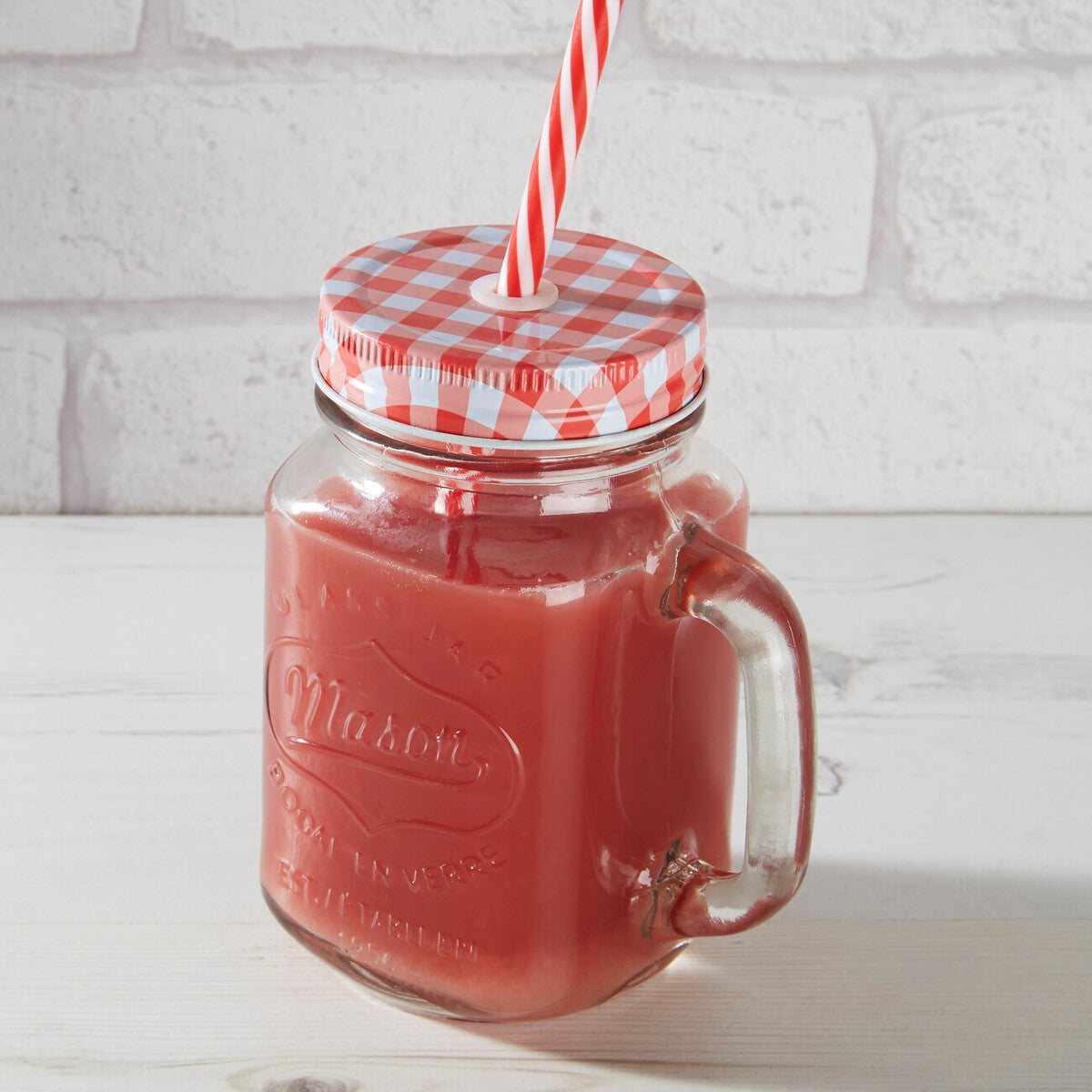 Glass mason jar with red liquid, checkered lid, and red straw against a white brick wall.