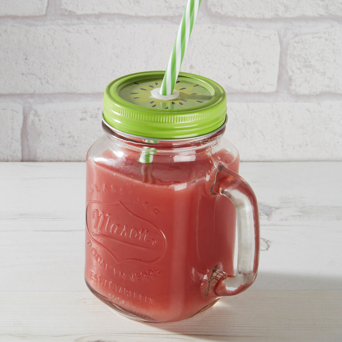 Red juice in a Mason jar with a green lid and straw against a white brick wall.