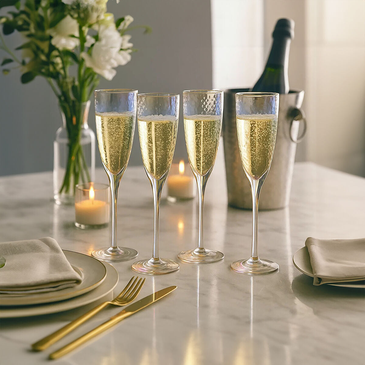 Four champagne glasses with bubbles on a table set for a meal, with a bottle of champagne and flowers in the background.