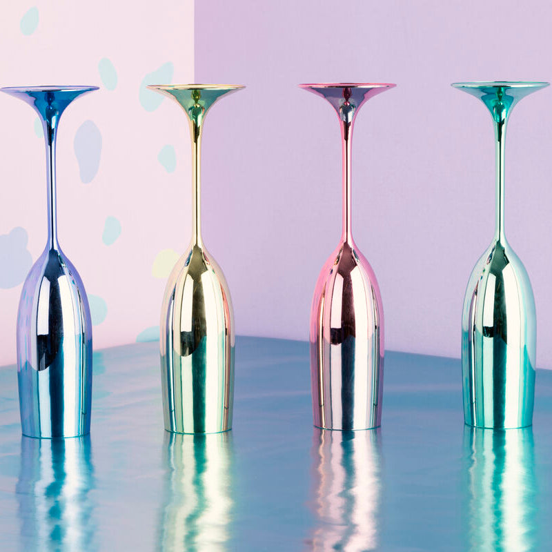 Four metallic wine glasses in different colors on a reflective surface with a colorful polka dot background.