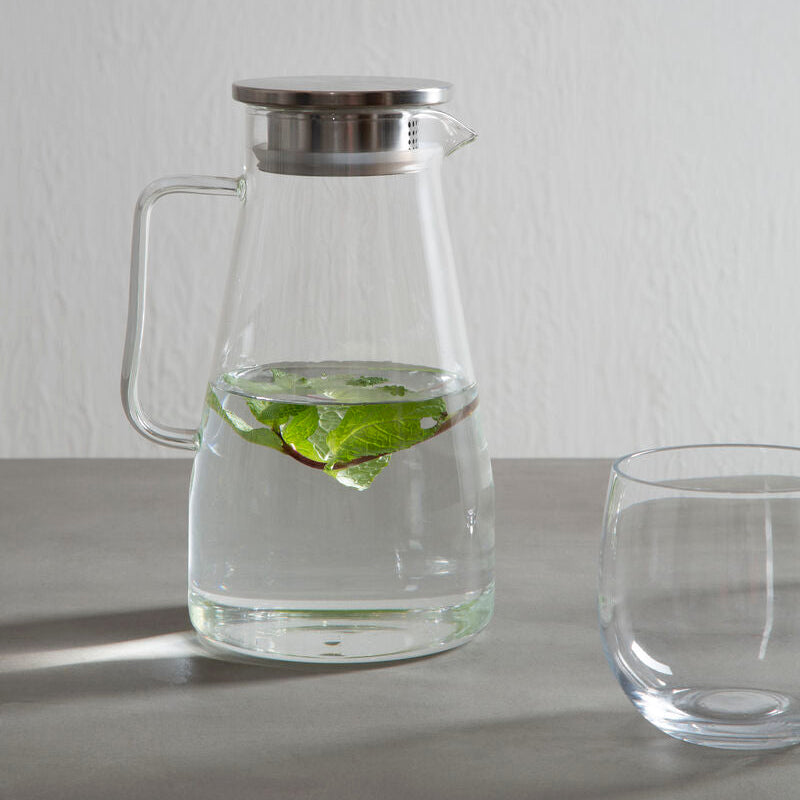 Clear glass pitcher with metal lid on a gray surface, filled with water and lime wedges.