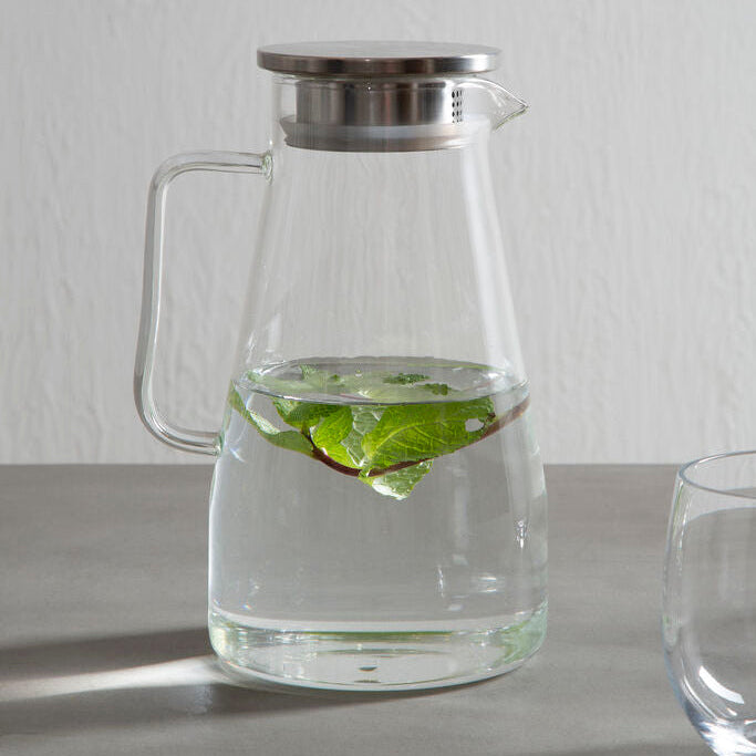 Clear glass pitcher with metal lid on a gray surface, filled with water and lime wedges.