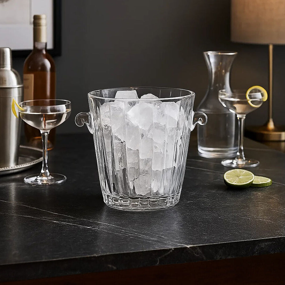 Glass ice bucket with ice cubes on a bar counter with cocktail-making equipment.