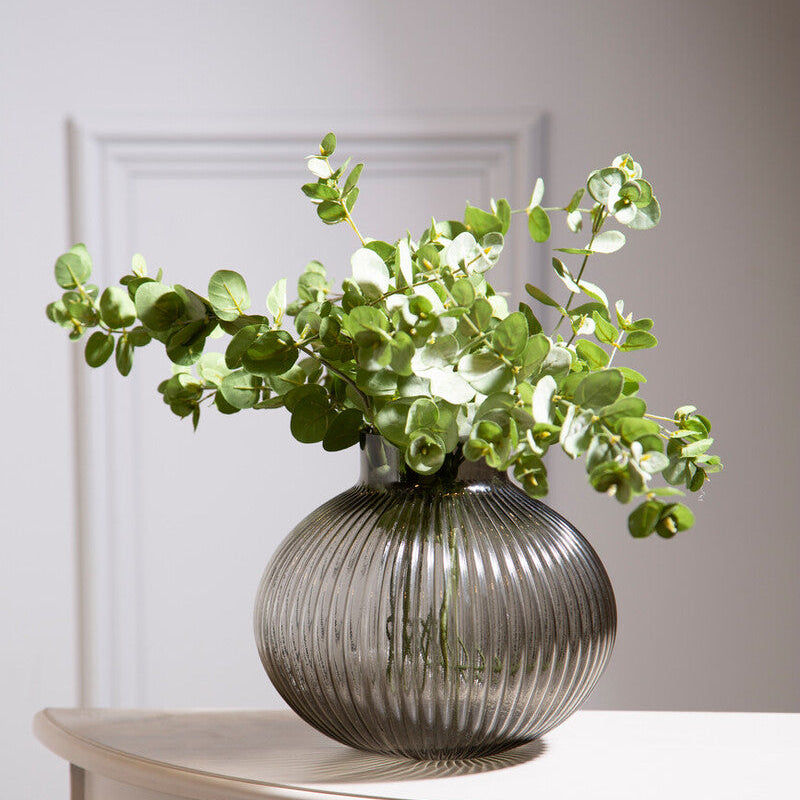 Glass vase with greenery on a wooden table against a white wall