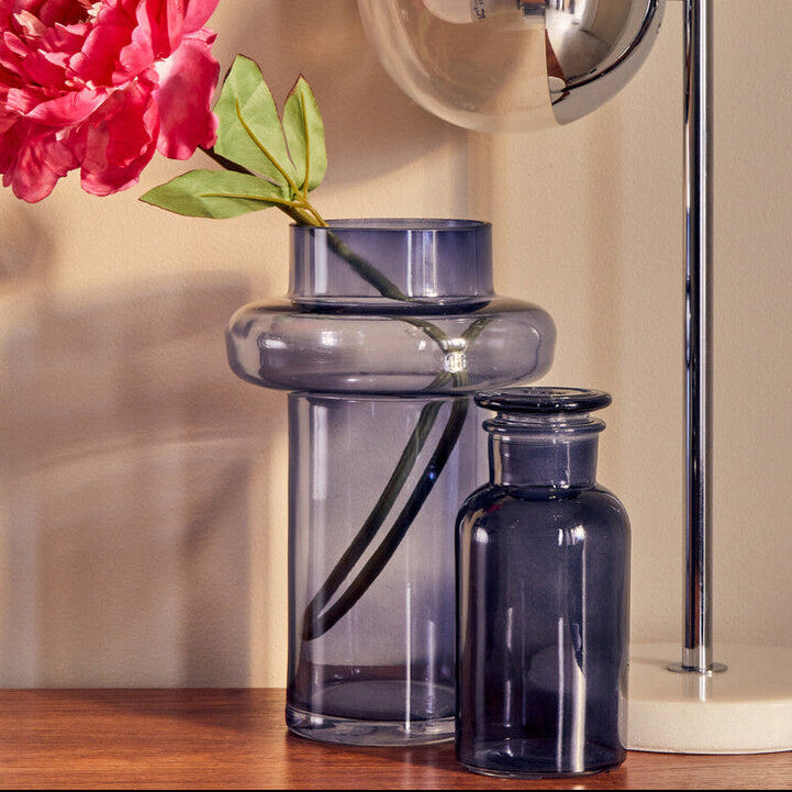 Glass vase with a pink flower on a wooden surface next to a lamp.