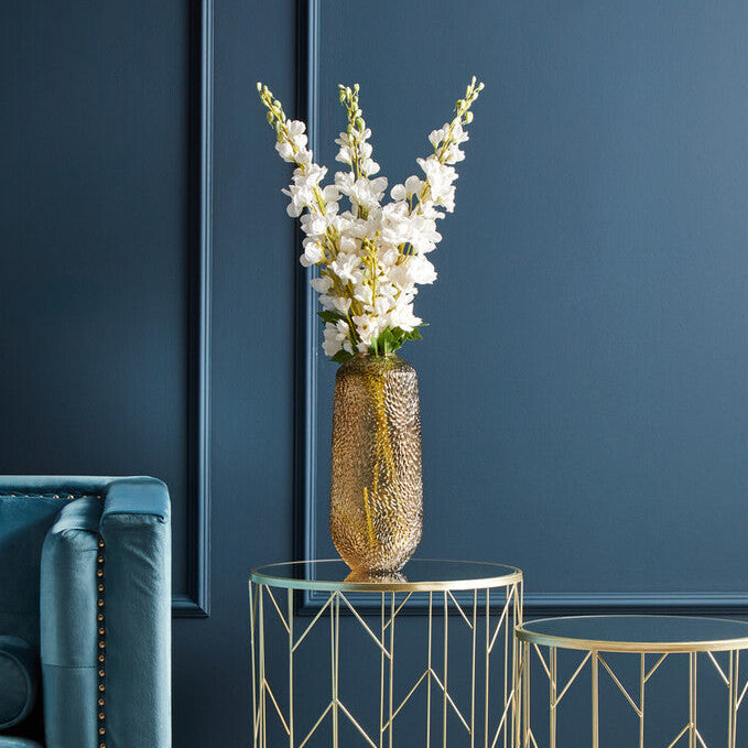 Gold side table with a vase of white flowers against a dark blue wall.