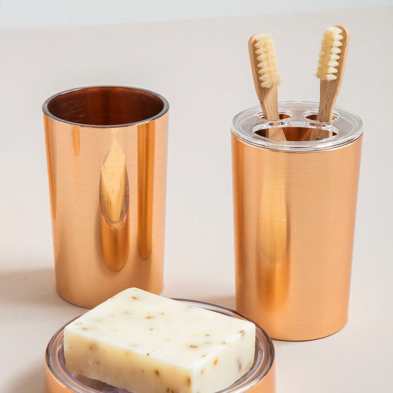 Set of bronze bathroom accessories including a soap dish, two cups with toothbrushes, and a bar of soap on a light background.